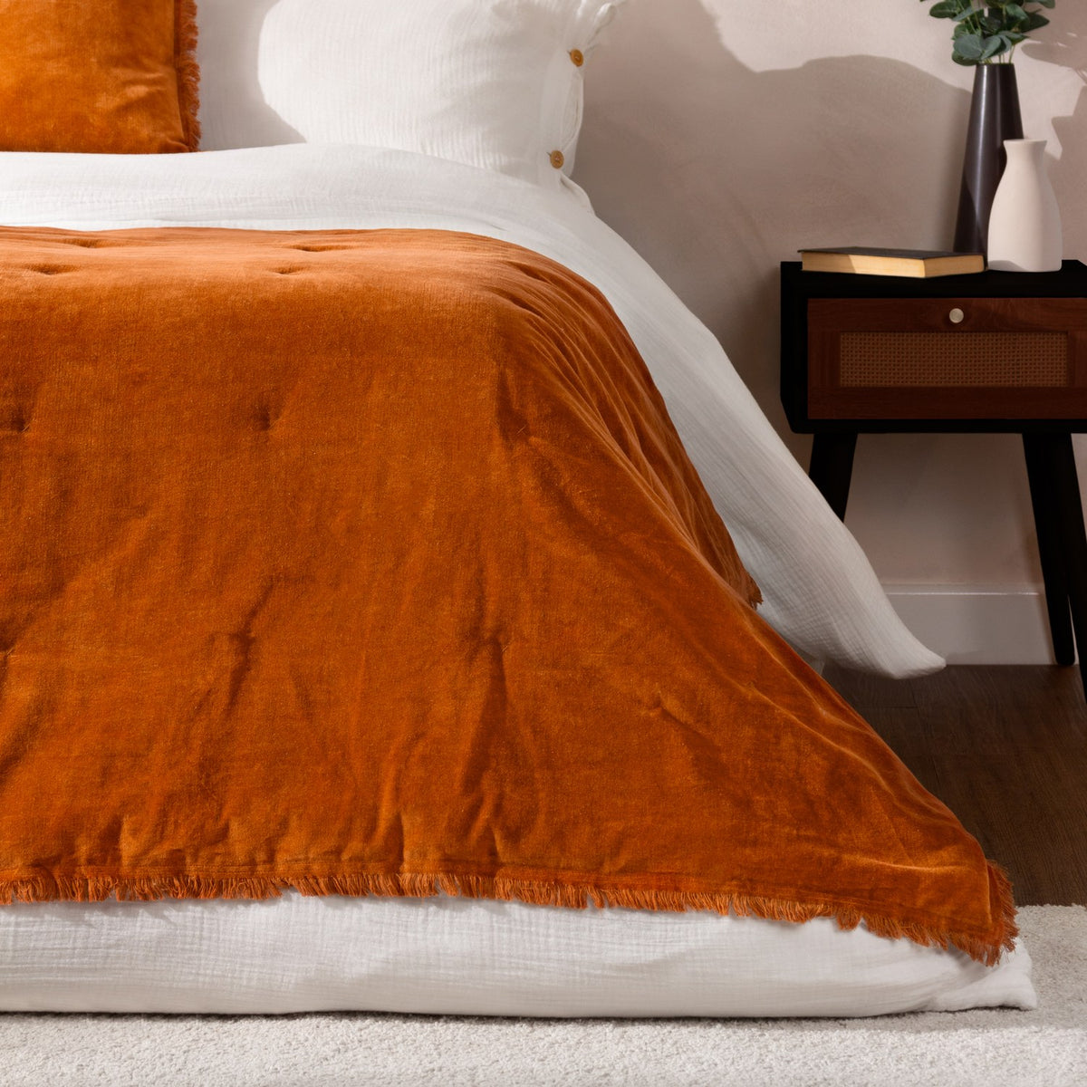 Jaye Velvet Cotton Bedspread in rust orange with fringed edge, shown on a bed with white bedding beside a dark wooden nightstand and vase.