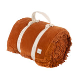 Jaye Velvet Cotton Bedspread in rust, rolled with fringed trim visible and secured by cream ties with a carry handle, shown on a plain white background.