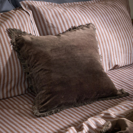 Jaye Velvet Cotton Cushion in bitter chocolate brown with fringed edges, shown on a bed with pink and white striped pillowcases and bedding.