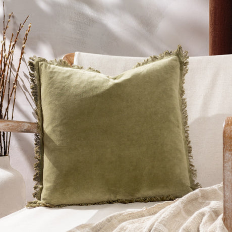 Jaye Velvet Cotton Cushion in moss green with fringed edges on all four sides, shown on a light sofa beside a neutral throw and dried branches.