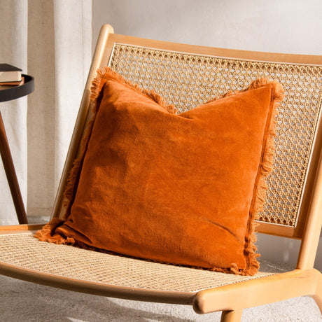 Jaye Velvet Cotton Cushion in rust orange with fringed edge trim, shown on a rattan and wood chair in a living room setting.