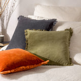 Jaye Velvet Cotton Cushion in rust orange with fringed trim edging, shown in front alongside olive green and charcoal cushions on a bouclé bed.