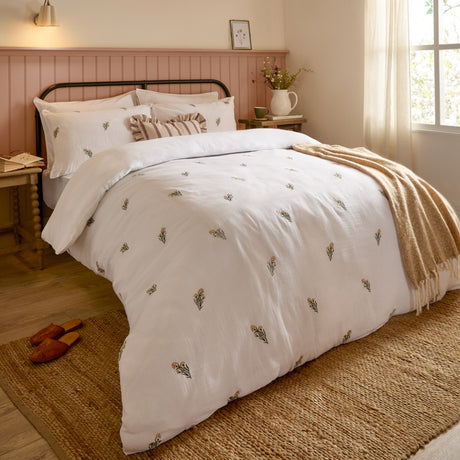 Josephine Cotton Floral Duvet Cover Set in white, styled on a bed with matching pillowcases, a striped cushion, and a beige throw in a pink-panelled bedroom.
