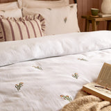 Josephine Cotton Floral Duvet Cover Set in white with pink-toned embroidered flowers, shown on a bed with matching pillowcases and striped cushions in a bedroom setting.