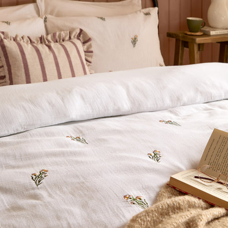 Josephine Cotton Floral Duvet Cover Set in white with pink-toned embroidered flowers, shown on a bed with matching pillowcases and striped cushions in a bedroom setting.