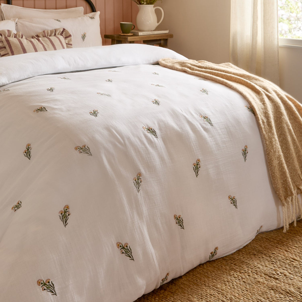 Josephine Cotton Floral Duvet Cover Set in white with embroidered peach and green floral sprigs, shown on a bed with a mustard throw and striped cushions in a bedroom setting.