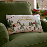 Rectangular Joy To All cushion on a green velvet armchair, cream front embroidered with a festive skating scene, gold "Merry Christmas" script, and a berry red piped edge.