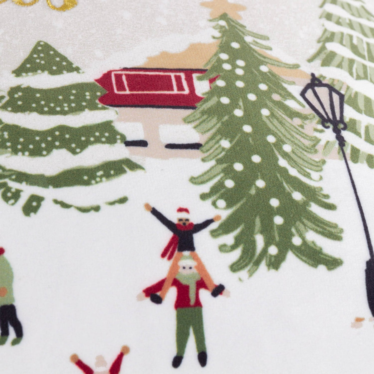 Close-up of the Joy To All velvet cushion showing an embroidered snowy village scene with a red cabin, green pine trees, a lamppost, and figures playing in festive winter clothing on a cream ground.