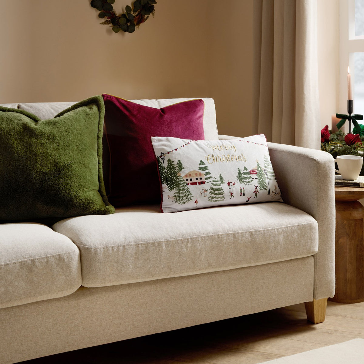 Joy To All rectangular cushion with cream base, embroidered festive village scene and gold "Merry Christmas" text, berry red piped edge, shown on a sofa with green and berry velvet cushions.