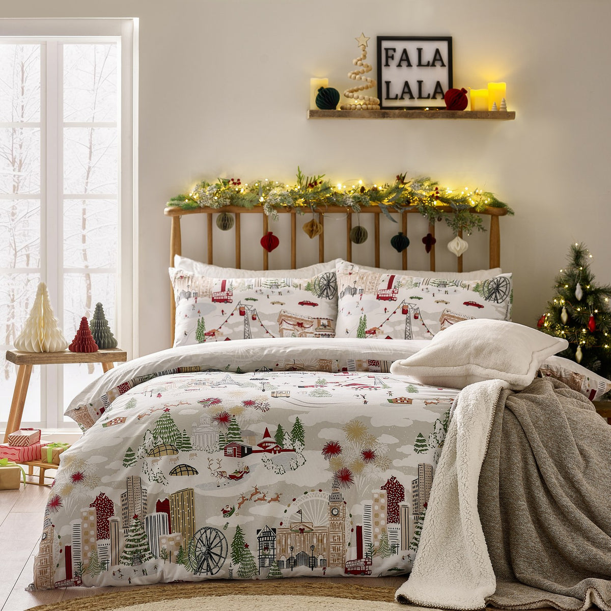 Joy To All duvet cover set on a bed, featuring a festive cityscape print with skyscrapers, a big wheel, cable cars and a sleigh in cream, grey, red and green, with matching pillowcases.