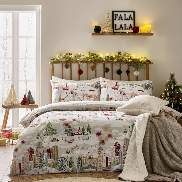 Joy To All duvet cover set on a bed, featuring a festive cityscape print with skyscrapers, a big wheel, cable cars and a sleigh in cream, grey, red and green, with matching pillowcases.