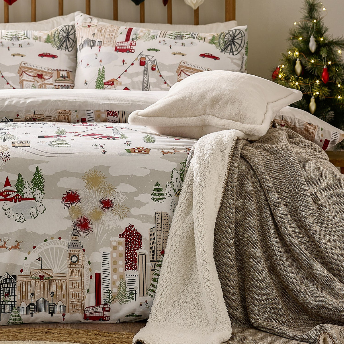 Joy To All reversible duvet cover set on a bed with matching pillowcases, showing a printed festive cityscape in cream, grey, red and green with a sherpa throw and cushion, near a lit Christmas tree.