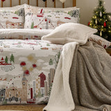 Joy To All reversible duvet cover set on a bed with matching pillowcases, showing a printed festive cityscape in cream, grey, red and green with a sherpa throw and cushion, near a lit Christmas tree.