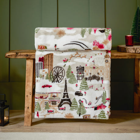 Joy To All cream polyester fleece throw, folded on a wooden bench, printed with a festive city scene of skylines, trees, cars and skaters in red, green and brown.