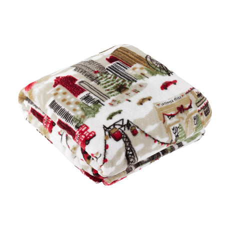 Folded Joy To All printed polyester throw in cream, featuring a festive scene of city buildings, trees, cable cars and taxis in red, green and brown, shown on a plain white background.