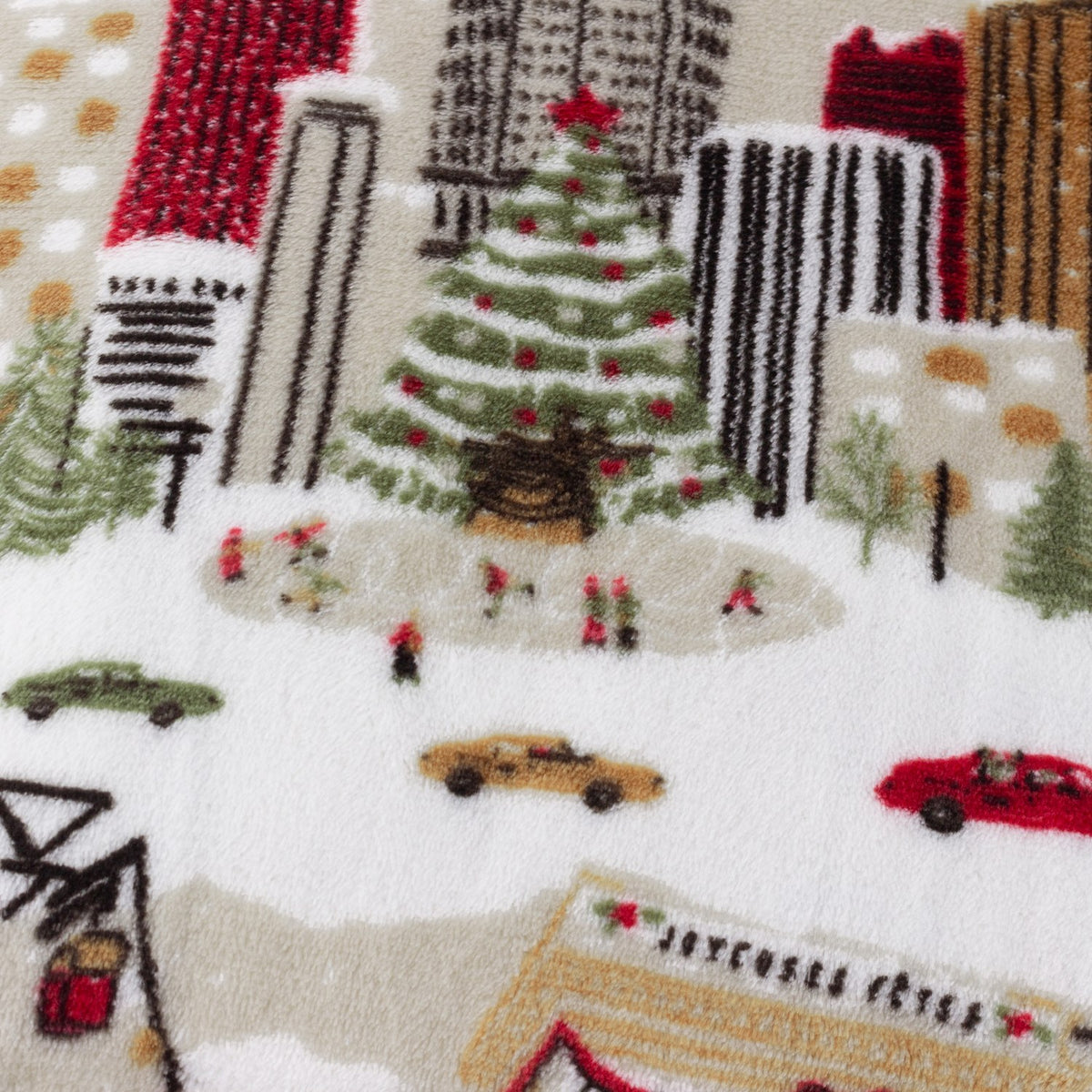 Close-up of the Joy To All printed polyester fleece throw in cream, showing a festive city scene with Christmas tree, cars, cable car and skaters in red, green and brown tones.