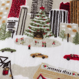 Close-up of the Joy To All printed polyester fleece throw in cream, showing a festive city scene with Christmas tree, cars, cable car and skaters in red, green and brown tones.