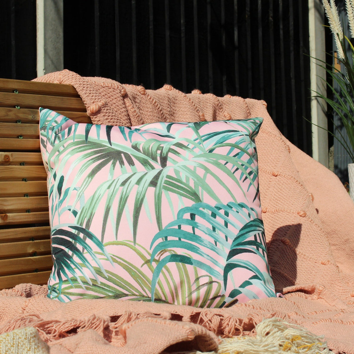 Jungle Reversible Polyester Cushion in Blush/Forest, square with green palm leaf print on a pink blush ground, shown on a wooden bench with a peach textured throw outdoors.