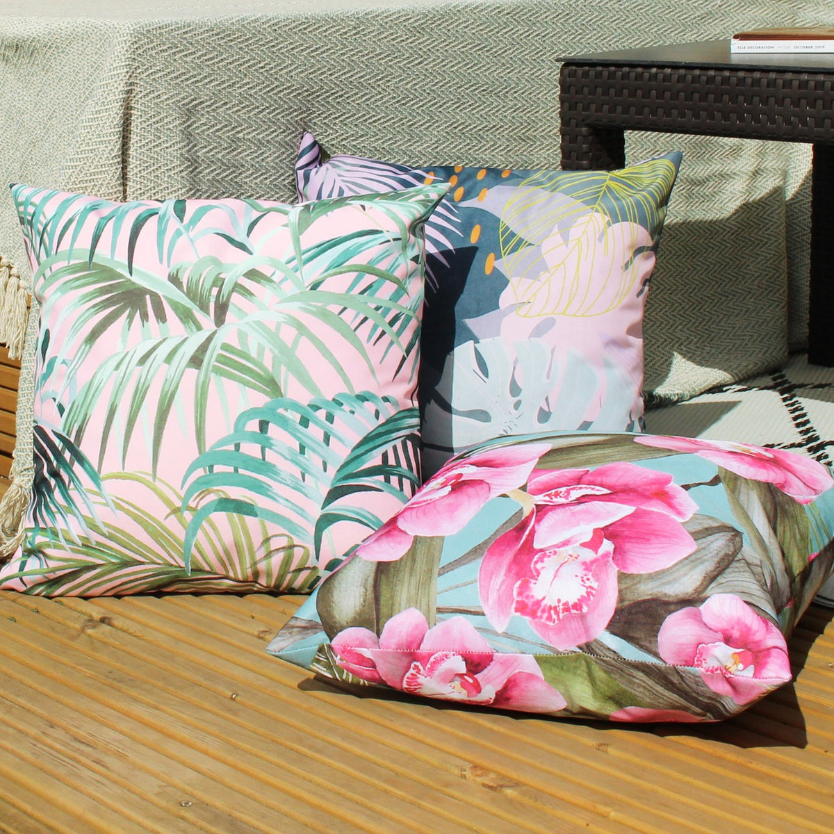 Jungle Reversible Polyester Cushion in Blush/Forest with palm frond print, shown outdoors on a wooden deck alongside floral and abstract patterned cushions.