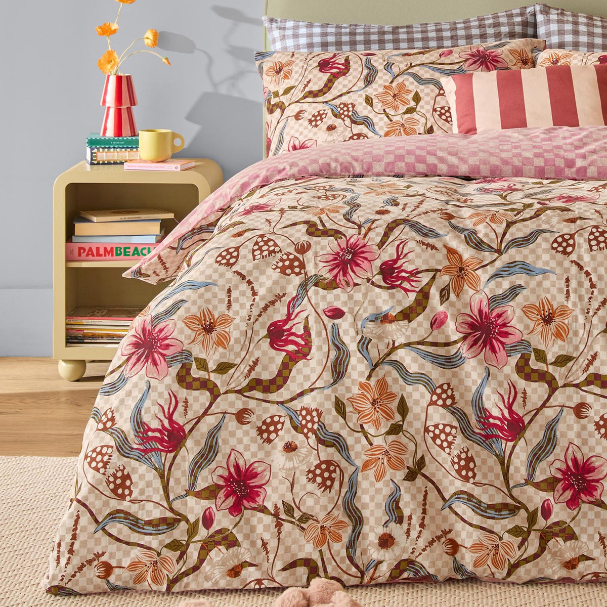 Juno Reversible Check Duvet Cover Set in a bedroom setting, floral pink, coral and rust print on a check ground, styled with striped and gingham cushions on a bed beside a yellow nightstand.