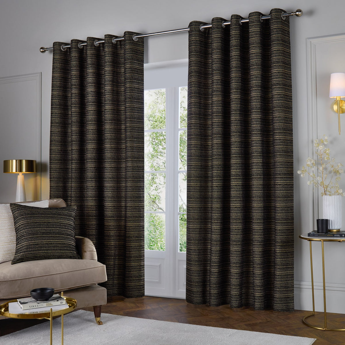 Jupiter Chenille Fully Lined Eyelet Curtains in Black/Gold, shown floor-length on a chrome pole in a living room with striped horizontal texture and a matching cushion nearby.