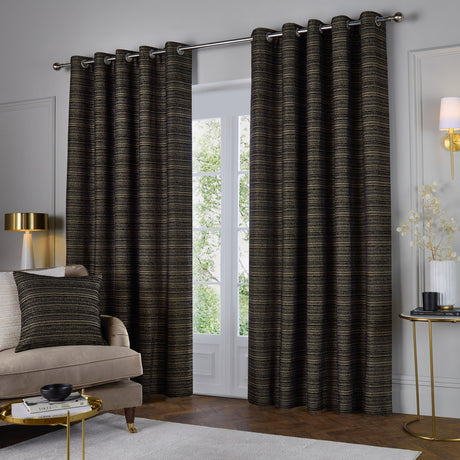 Jupiter Chenille Fully Lined Eyelet Curtains in Black/Gold, shown floor-length on a chrome pole in a living room with striped horizontal texture and a matching cushion nearby.