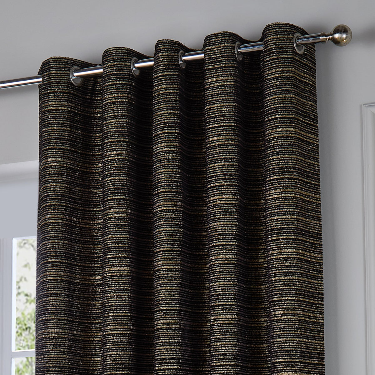 Jupiter Chenille Fully Lined Eyelet Curtain in black with fine gold horizontal striping, shown hung on a chrome pole with eyelet header in a bright room.