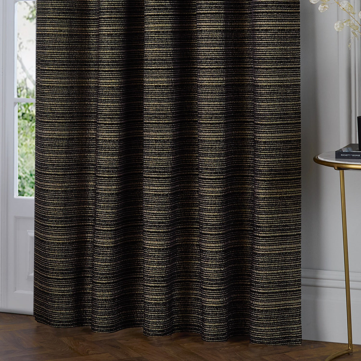Black and gold Jupiter chenille fully lined eyelet curtains with horizontal gold threading, hanging beside a French door and side table in a living room.