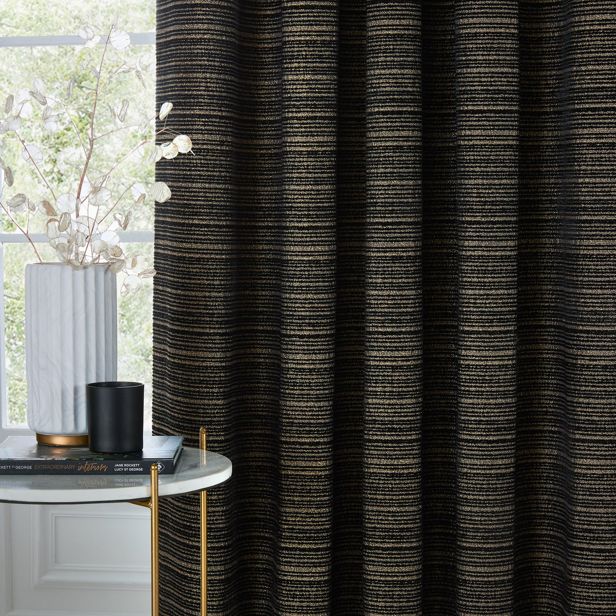 Jupiter Chenille Fully Lined Eyelet Curtains in Black/Gold, showing fine gold pearlescent stripes on a black chenille jacquard fabric, styled in a living room with a marble side table and vase.