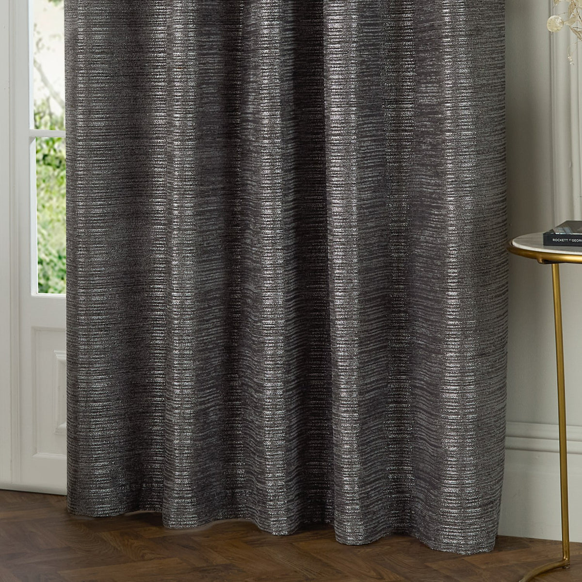 Jupiter Chenille Fully Lined Eyelet Curtains in graphite, showing a textured woven fabric with fine horizontal lines, hung beside a window in a living room setting.