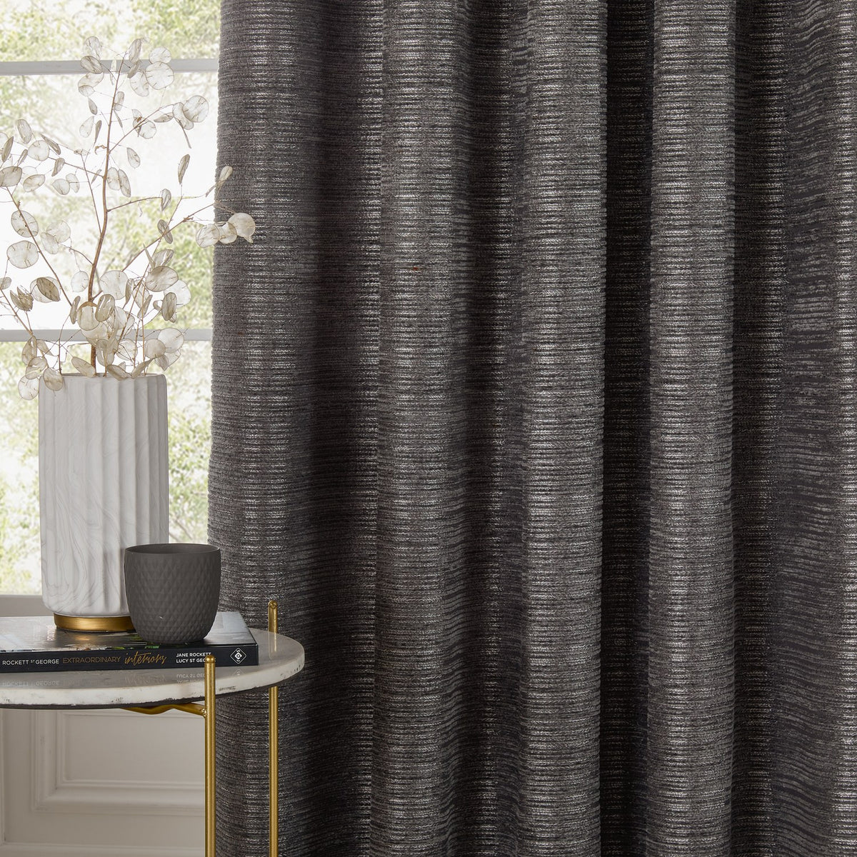 Jupiter Chenille Fully Lined Eyelet Curtains in graphite, textured fabric with fine horizontal lines, shown fully drawn beside a window in a living room setting.