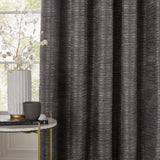 Jupiter Chenille Fully Lined Eyelet Curtains in graphite, textured fabric with fine horizontal lines, shown fully drawn beside a window in a living room setting.