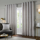Jupiter Chenille Fully Lined Eyelet Curtains in silver, shown as a pair with a fine horizontal woven texture, hung on a chrome pole in a living room beside a glass door.