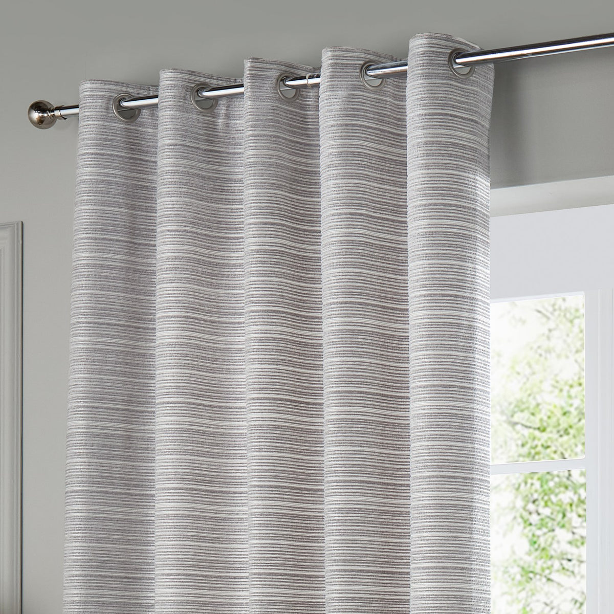 Jupiter Chenille Fully Lined Eyelet Curtains in silver, hung on a chrome pole with metal grommets, showing a fine horizontal woven texture beside a window.