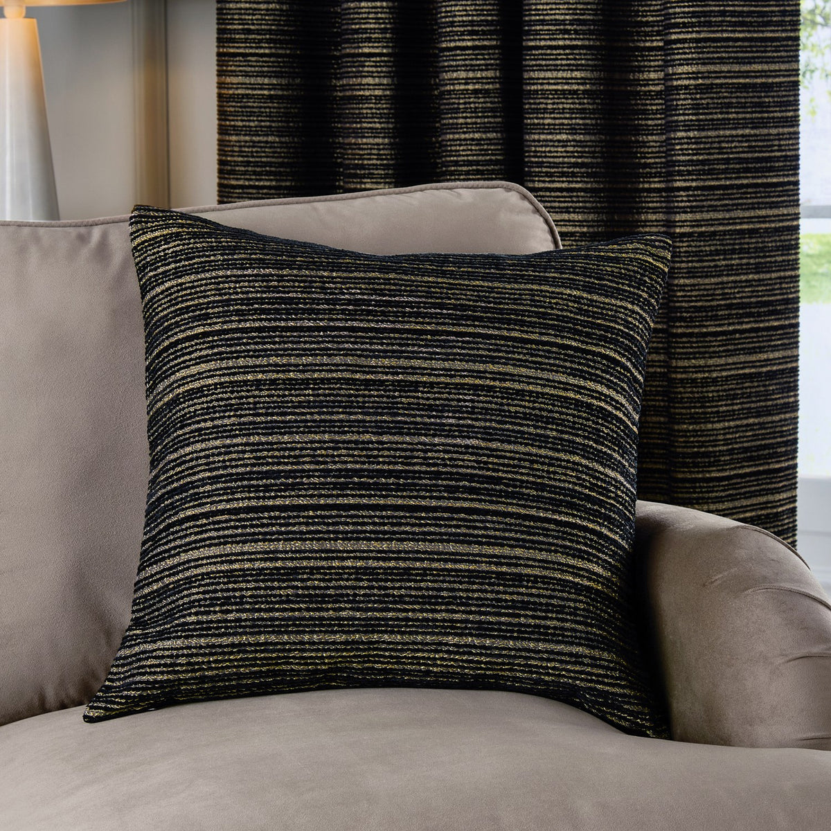 Jupiter Jacquard Chenille Cushion in black with fine gold horizontal stripes, styled on a taupe armchair against a matching striped curtain in a living room setting.