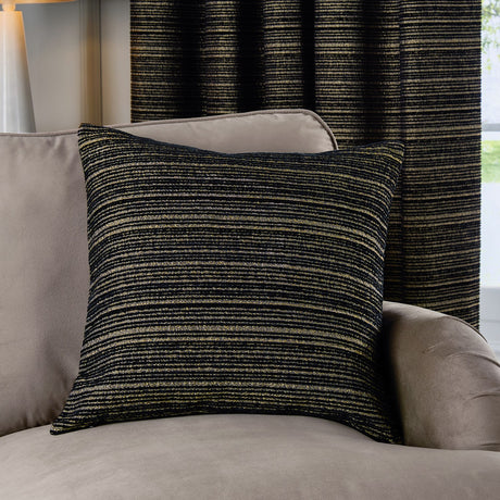 Jupiter Jacquard Chenille Cushion in black with fine gold horizontal stripes, styled on a taupe armchair against a matching striped curtain in a living room setting.