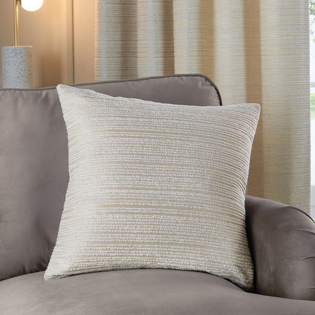 Jupiter Jacquard Chenille Cushion in ivory with subtle gold ridged texture, styled on a grey armchair against curtains in a living room setting.
