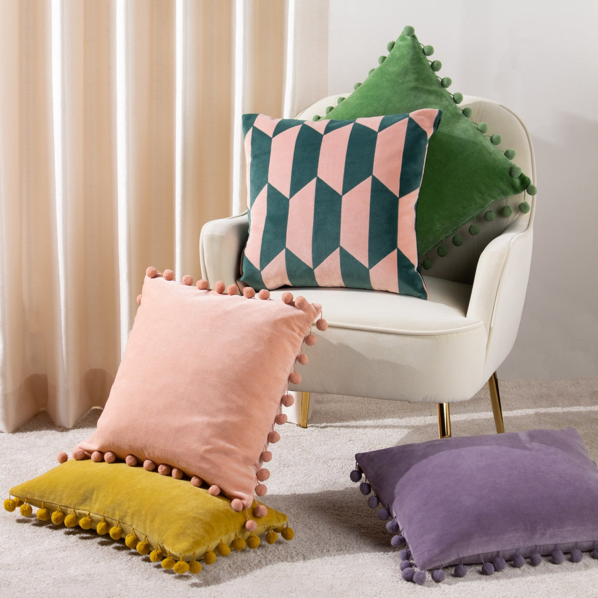 Kalho Jacquard Velvet Cushion in pink and green geometric pattern, styled on a cream armchair with pink, yellow, and purple pom-pom trim cushions nearby.