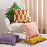 Kalho Jacquard Velvet Cushion in pink and ochre with a geometric block pattern, shown on an armchair alongside pom-pom trimmed cushions in green, purple, and yellow.