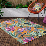 Kali Animals square cushion with orange, green, pink and purple jungle print of lemurs and toucans, shown on a matching patterned rug beside an orange rattan chair on a wooden deck.