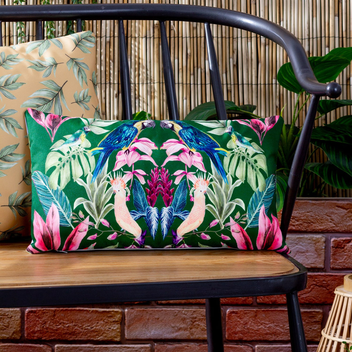 Kali Birds rectangular cushion in dark green polyester with a symmetrical parrot and cockatoo print, styled on a black metal bench beside a bamboo backdrop.
