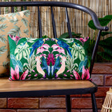 Kali Birds rectangular cushion in dark green polyester with a symmetrical parrot and cockatoo print, styled on a black metal bench beside a bamboo backdrop.