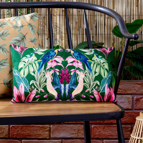 Kali Birds rectangular cushion in dark green polyester with a symmetrical parrot and cockatoo print, styled on a black metal bench beside a bamboo backdrop.
