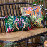 Kali Birds rectangular cushion in dark green with symmetrical parrot and tropical bird print, shown on a bench beside an orange jungle-print cushion.