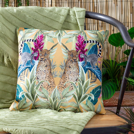 Kali Leopards square cushion in amber, green and pink, printed with mirrored leopards, lemurs and jungle foliage, shown outdoors on a chair with a green throw.