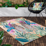 Kali Leopards reversible polyester cushion in multicolour, printed with mirrored leopards, lemurs and jungle foliage on an amber ground, shown on a black outdoor chair beside a matching rug.