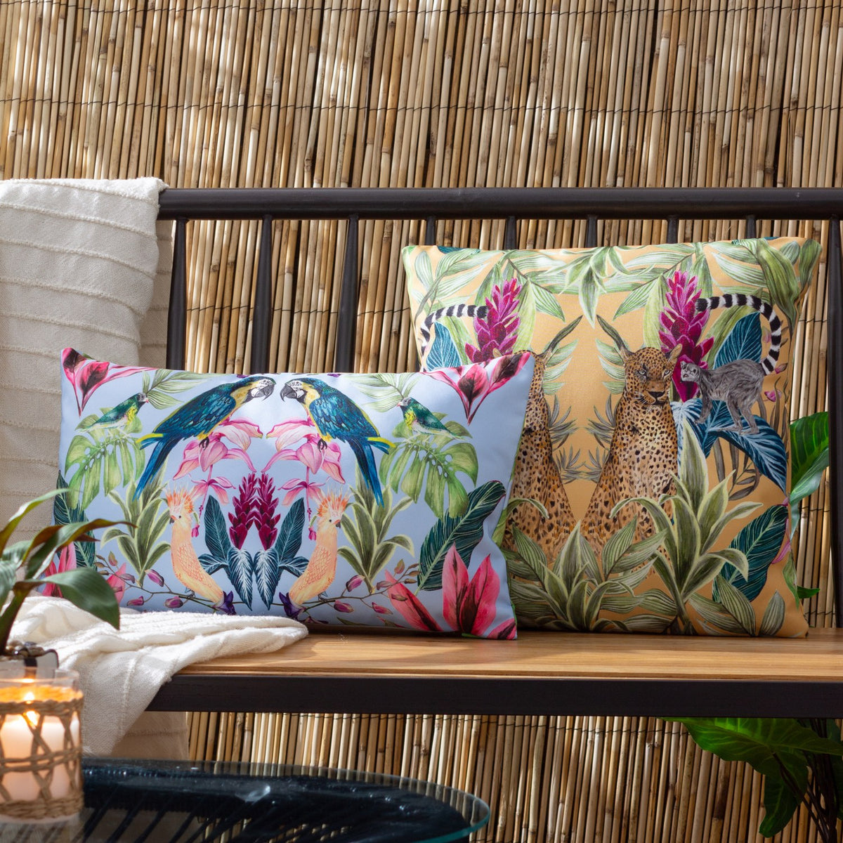 Kali Leopards square cushion in amber with mirrored leopard and lemur jungle print, shown beside a bird-print cushion on an outdoor bench.