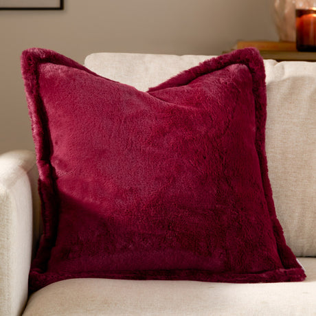 Kallu Plain Faux Fur Cushion in deep berry red with an oxford border, shown on a light beige sofa in a living room setting.