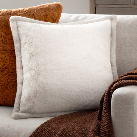 Kallu Reversible Faux Fur Cushion in ecru with Oxford flange border, shown on a sofa alongside a burnt orange cushion and brown throw.