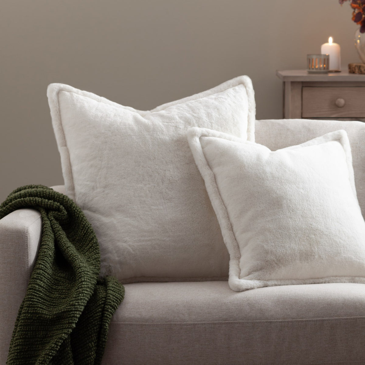 Two Kallu reversible faux fur cushions in ecru with Oxford flange borders, styled on a beige sofa beside a green throw and lit side table.